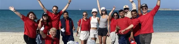A group of cheerful volunteers wearing red and white attire celebrate on a sunny beach, with blue water and clear skies in the background.
