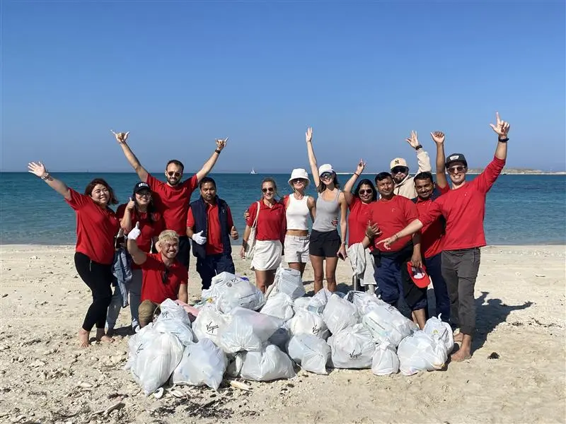 A group of cheerful volunteers wearing red and white attire celebrate on a sunny beach, with blue water and clear skies in the background.