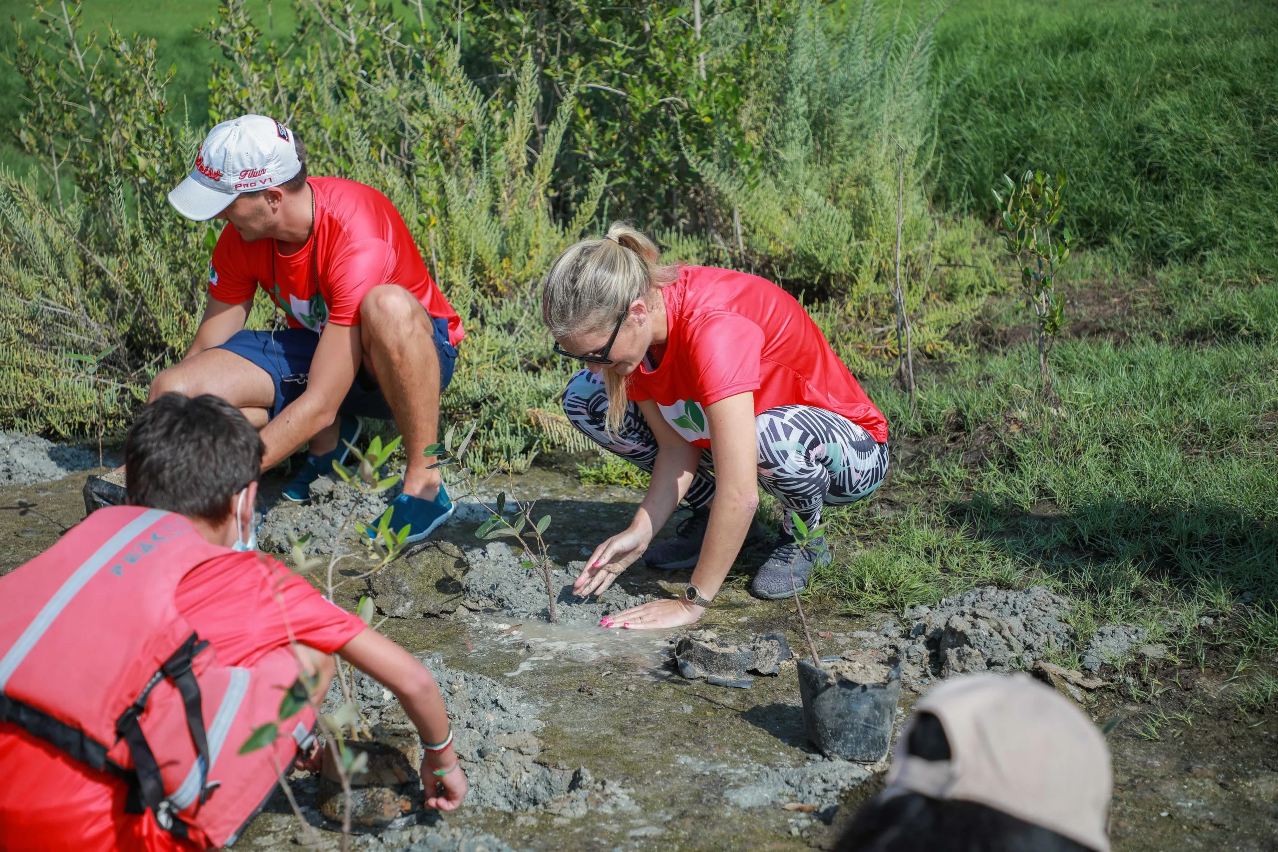 People in red shirts planting by a stream, surrounded by greenery.