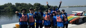 A group of happy kayakers in life jackets ready to paddle, near colorful kayaks by a lake.