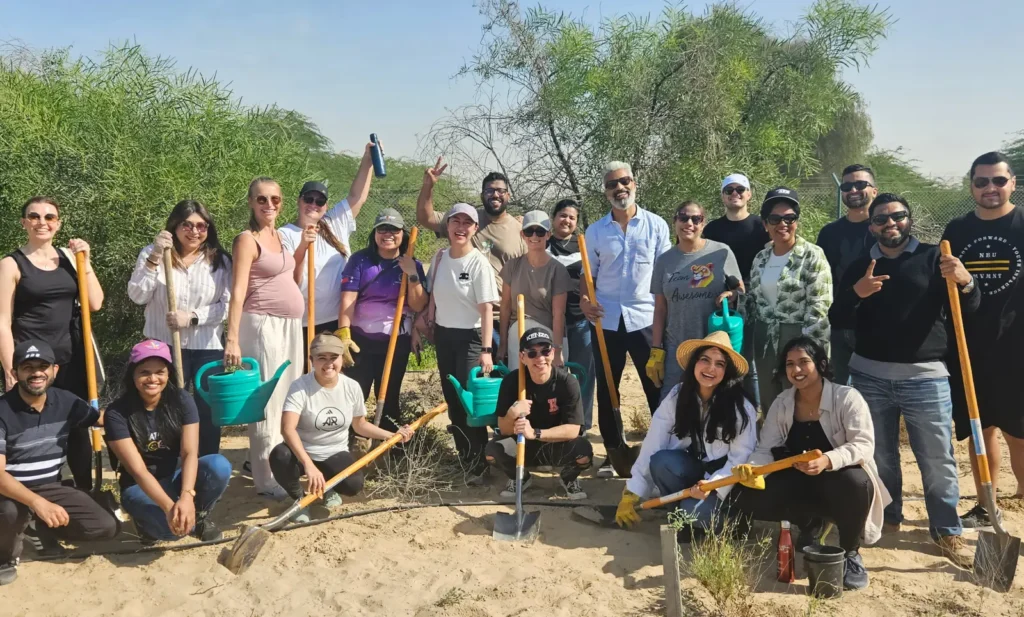 Group of people smiling outdoors, some holding tools, with greenery in the background.
