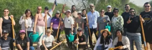 Group of people smiling outdoors, some holding tools, with greenery in the background.