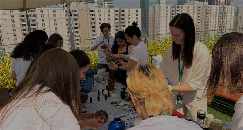 A group of people engaged in a lively outdoor gathering, interacting around a table with various items. Lush greenery in the background.