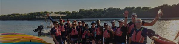 A group of excited individuals in life vests pose together near a body of water, with kayaks and lush greenery in the background.