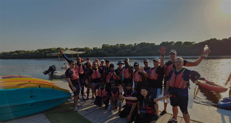 A group of excited individuals in life vests pose together near a body of water, with kayaks and lush greenery in the background.