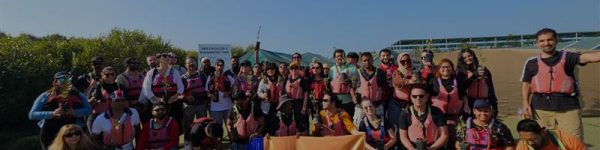 A large group of people wearing life jackets gathers outdoors, smiling and posing for a photo under clear blue skies.