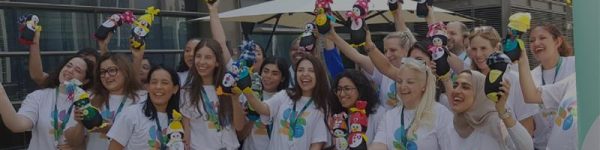 A group of individuals in white t-shirts joyfully holds up colorful plush toys, celebrating a lively event outdoors.