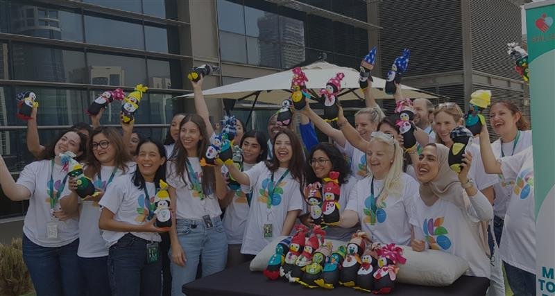 A group of individuals in white t-shirts joyfully holds up colorful plush toys, celebrating a lively event outdoors.