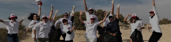 A group of enthusiastic volunteers in matching t-shirts and hats celebrating while cleaning up a sandy beach environment.