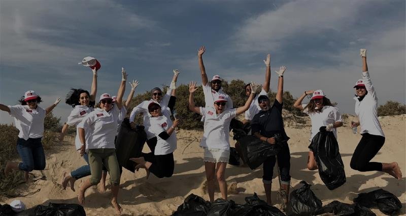 A group of enthusiastic volunteers in matching t-shirts and hats celebrating while cleaning up a sandy beach environment.