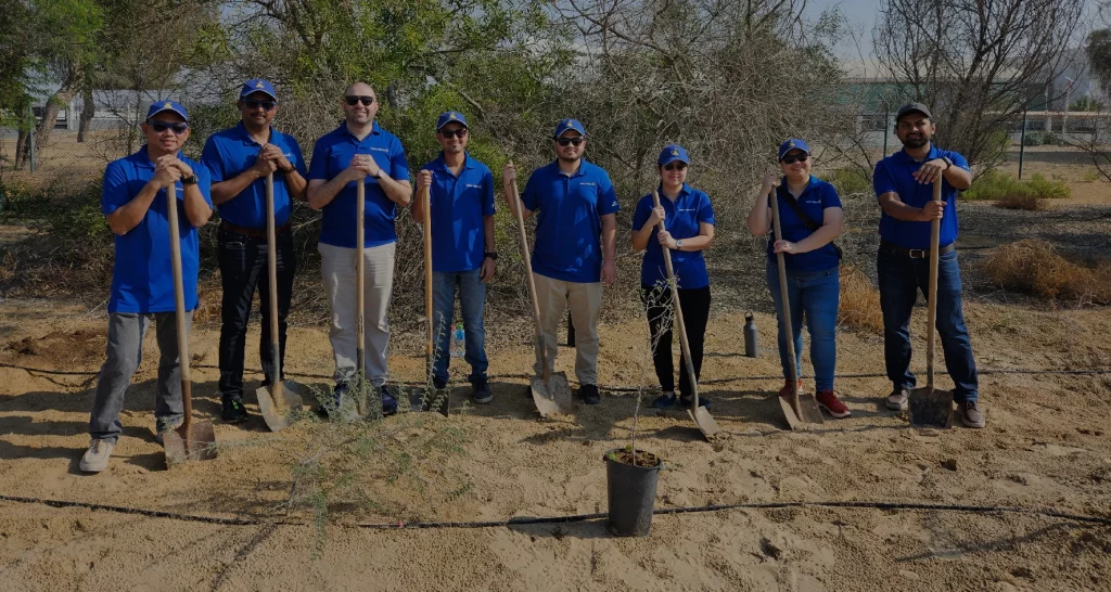 A group of six individuals in blue shirts holding shovels, standing on sandy ground, ready for a groundbreaking event.