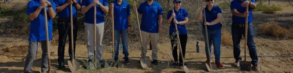 A group of six individuals in blue shirts holding shovels, standing on sandy ground, ready for a groundbreaking event.