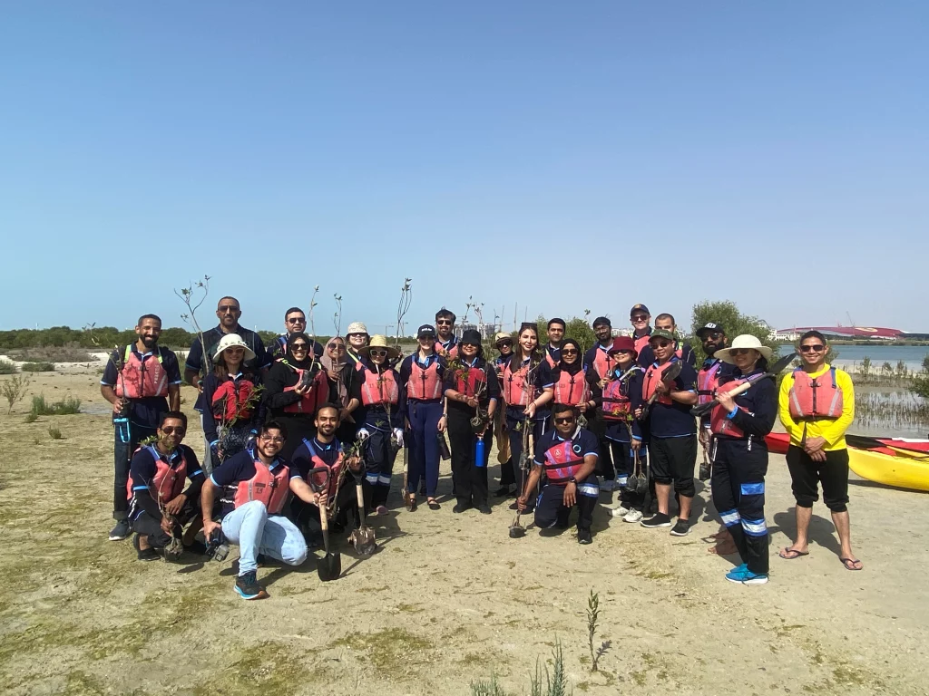 A group of environmental volunteers in life jackets poses with saplings near a waterway, ready for a tree-planting initiative.