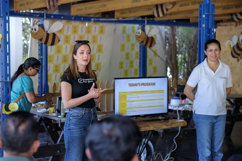 Two women stand beside a monitor displaying "Today's Program" at an event, with notes pinned on the wall behind them.