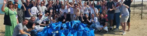 A large group of people gathered on the beach, smiling and waving, with a pile of blue bags in front of them, celebrating a clean-up event.