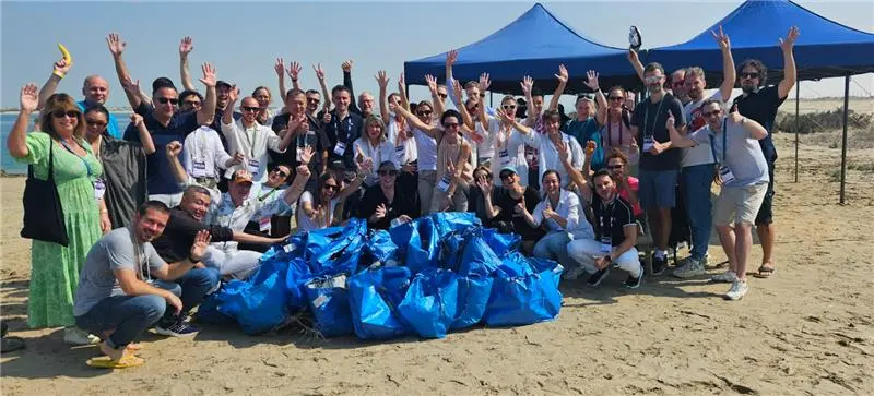 A large group of people gathered on the beach, smiling and waving, with a pile of blue bags in front of them, celebrating a clean-up event.