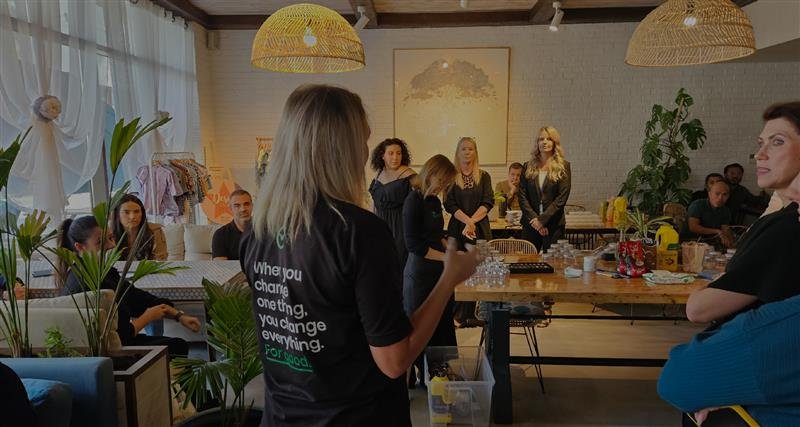 A group of people engaged in a workshop, with a presenter addressing attendees in a bright, modern environment filled with greenery.