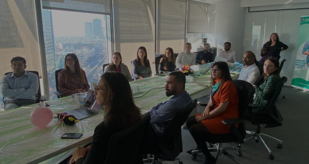 A diverse group of professionals sits at a conference table, engaged in discussion, with a city skyline visible through large windows.