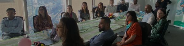 A diverse group of professionals sits at a conference table, engaged in discussion, with a city skyline visible through large windows.