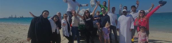 A diverse group of people joyfully celebrating on a sunny beach, with the sea and a clear blue sky in the background.