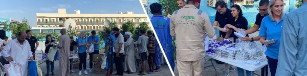 A community event with people in line receiving food and supplies from tables, set against a backdrop of a building and greenery.