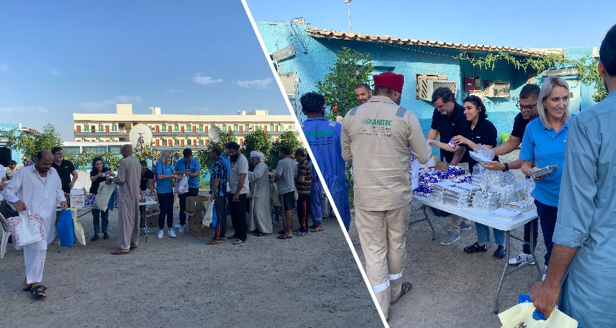 A community event with people in line receiving food and supplies from tables, set against a backdrop of a building and greenery.