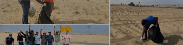 A group of volunteers collects trash in a desert area, smiling and posing for a photo while another volunteer cleans the sand.