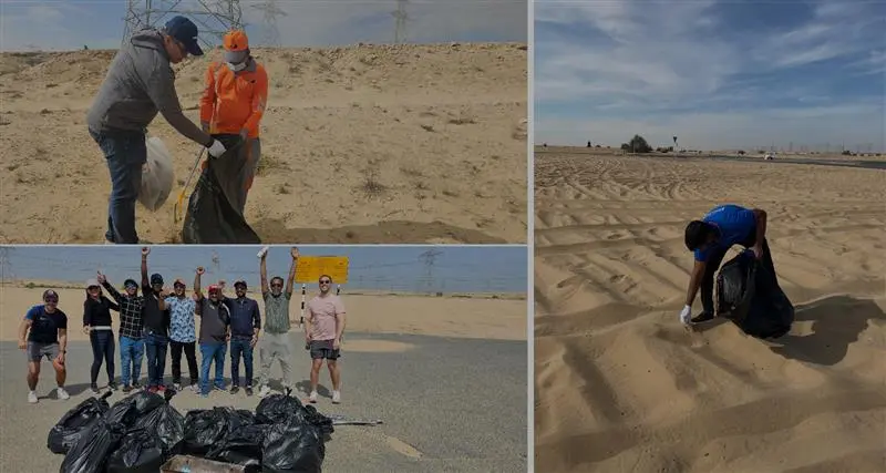A group of volunteers collects trash in a desert area, smiling and posing for a photo while another volunteer cleans the sand.