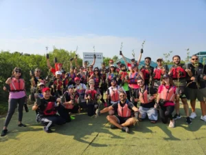 A diverse group of participants in life vests poses with small plants, celebrating a coastal restoration event under a clear blue sky.