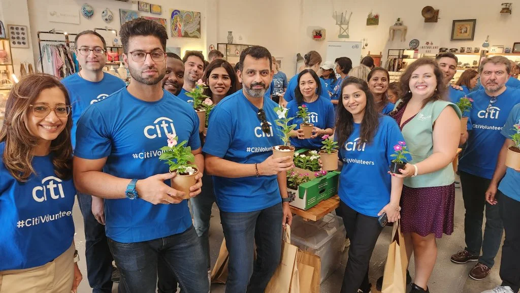 A group of volunteers in blue t-shirts hold potted plants while standing inside a vibrant indoor space filled with decorations.
