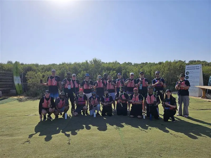 A group of 22 individuals in pink life jackets pose together outdoors, surrounded by greenery and kayaks in the background.