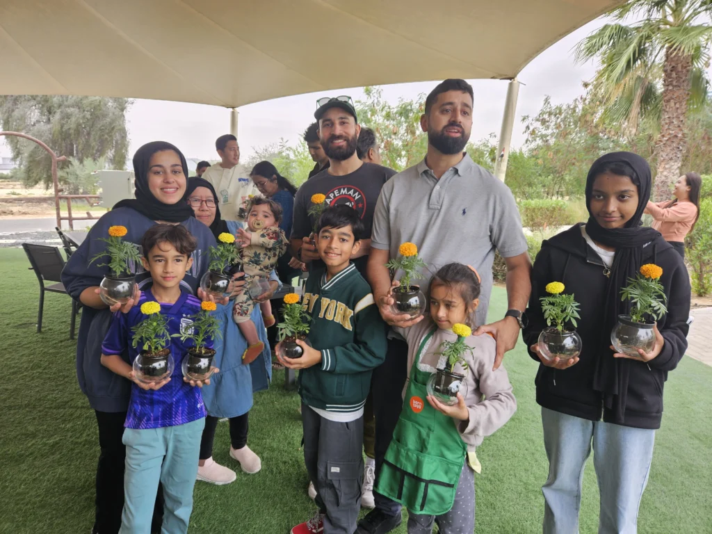 A group of individuals, including children, proudly hold small potted marigold plants in glass containers, smiling for the camera.