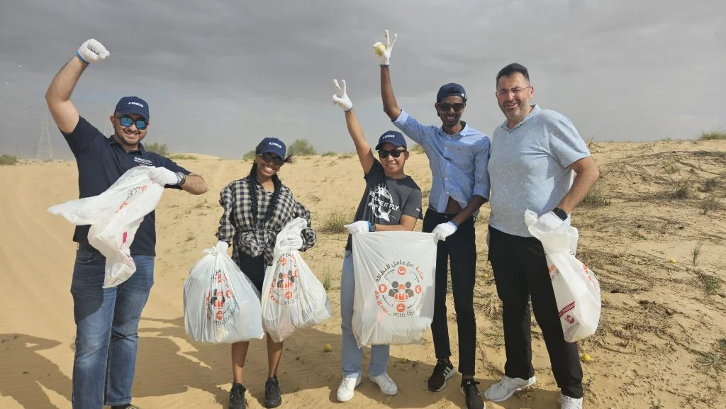A group of individuals stands on a sandy landscape, holding bags of collected litter and smiling, promoting environmental awareness.