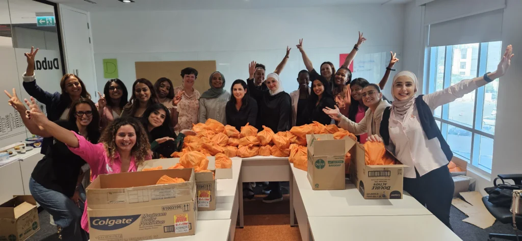 A diverse group of people celebrates around tables filled with neatly wrapped orange packages, showcasing teamwork and community spirit.