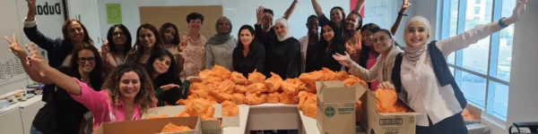 A diverse group of people celebrates around tables filled with neatly wrapped orange packages, showcasing teamwork and community spirit.