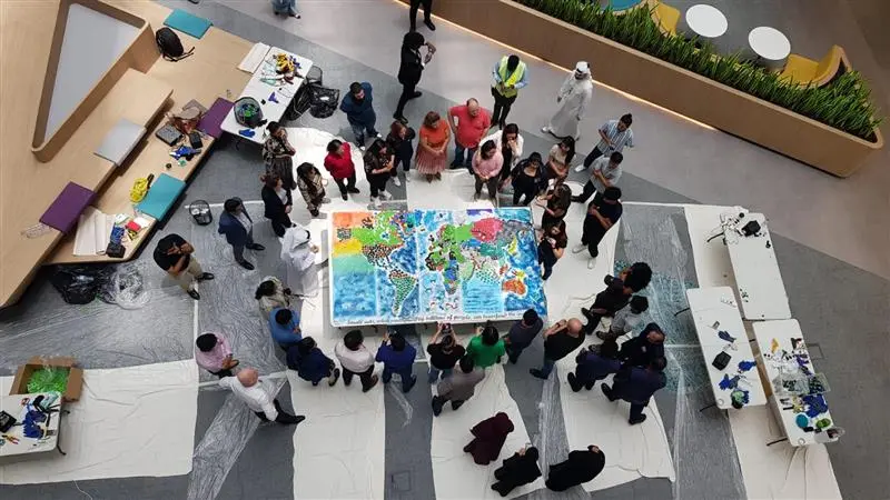 Aerial view of a diverse group of people gathered around a colorful, large world map painted on a table in a communal space.