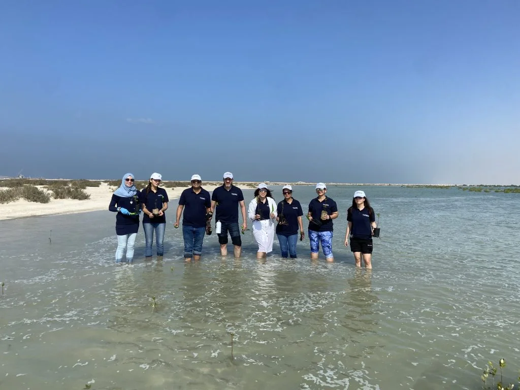 A group of individuals stands in shallow water, wearing navy shirts and hats, engaged in environmental conservation activities.