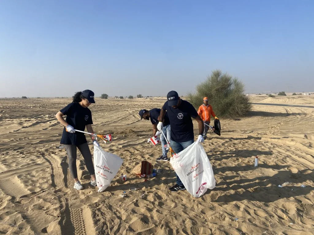 Volunteers in black t-shirts collect trash in the desert, using litter pickers and bags, surrounded by sandy dunes and sparse vegetation.