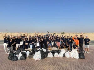 A large group of volunteers in matching shirts celebrates after a cleanup, surrounded by filled trash bags on a sunny day.
