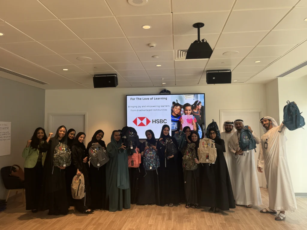 A group of individuals in traditional attire holds backpacks, with a presentation about educational support from HSBC in the background.