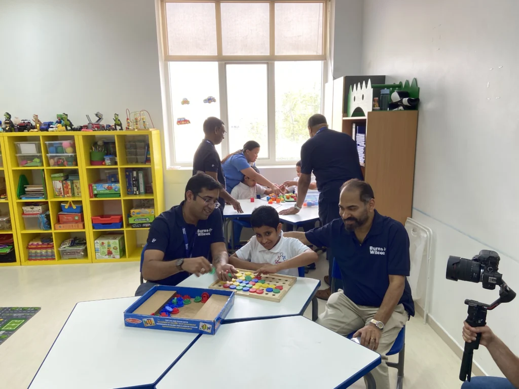 Two adults interact with a child at a table with colorful educational toys in a bright classroom, while others work in the background.
