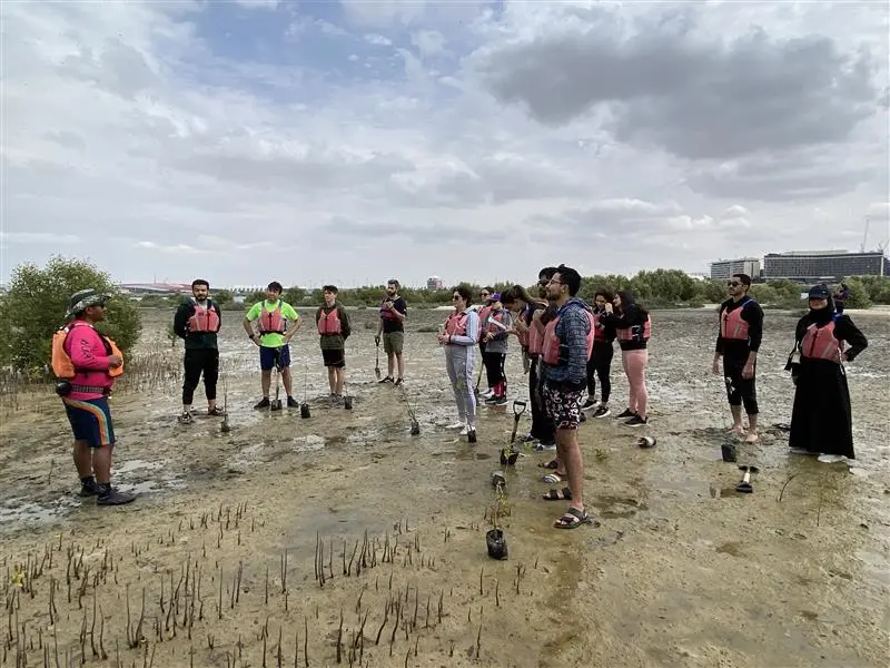 A diverse group of individuals wearing life jackets listens attentively to a guide in a muddy, natural setting, promoting environmental awareness.