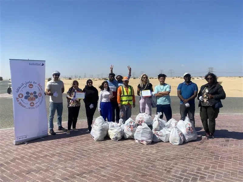 A diverse group of volunteers poses proudly holding certificates, with bags of collected waste in a desert landscape background.