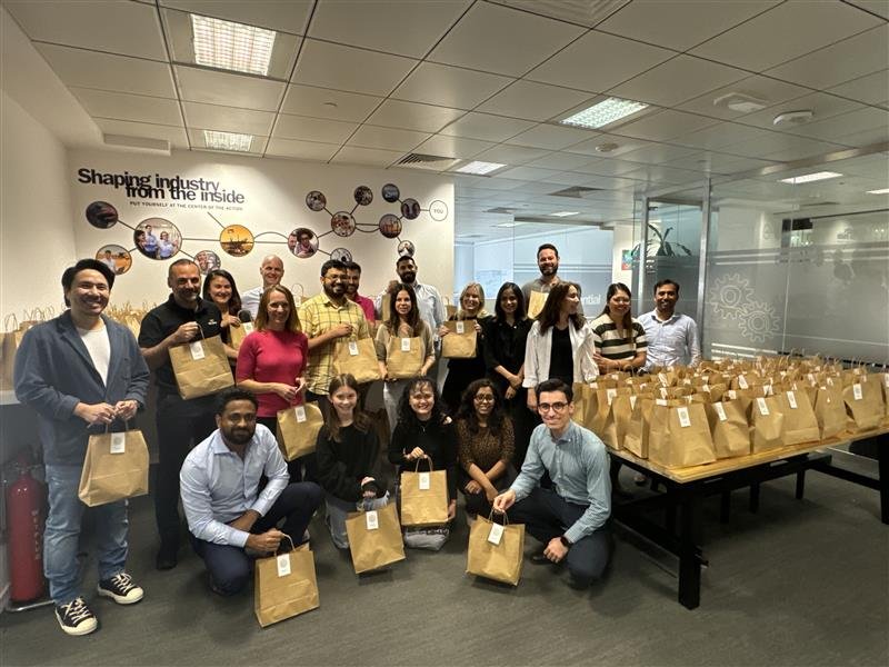 A diverse group of people smiling while holding brown paper bags in an office, with a table and floor filled with neatly arranged brown bags, indicating a team volunteer or community packaging activity.