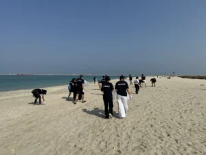 A group of volunteers in black t-shirts walk along a sandy beach, collecting litter near the calm turquoise waters.