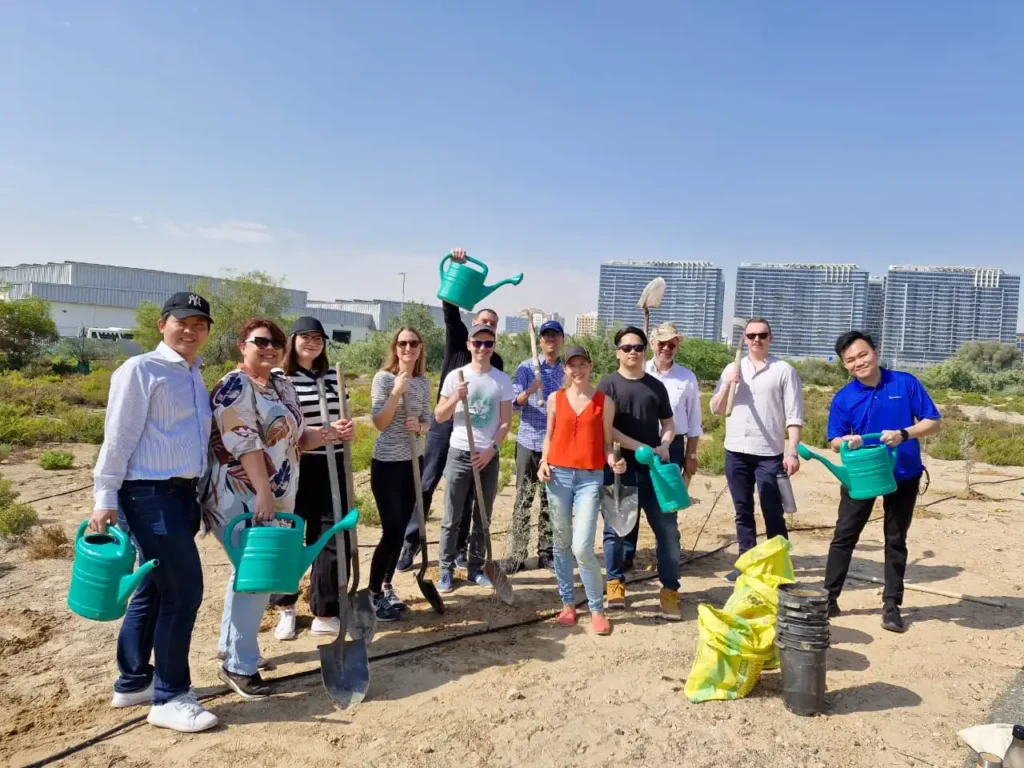 A group of people stands outdoors, smiling and holding gardening tools and watering cans, with modern buildings in the background.