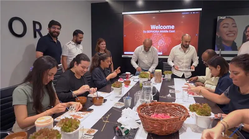 A group of people at a dining table engaged in conversation and dining, with plates and drinks present and a presentation screen in the background.