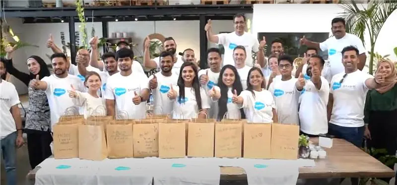 Group of volunteers wearing matching t-shirts giving thumbs up while standing behind eco-friendly paper bags during a community event.