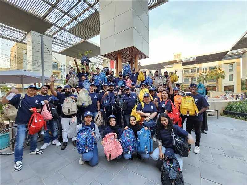 Large group of volunteers wearing matching shirts and caps, holding colorful backpacks during an outdoor community outreach event