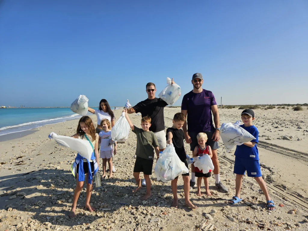 A group of adults and children stands on a sandy beach, holding bags of collected litter, with a clear blue sea in the background.